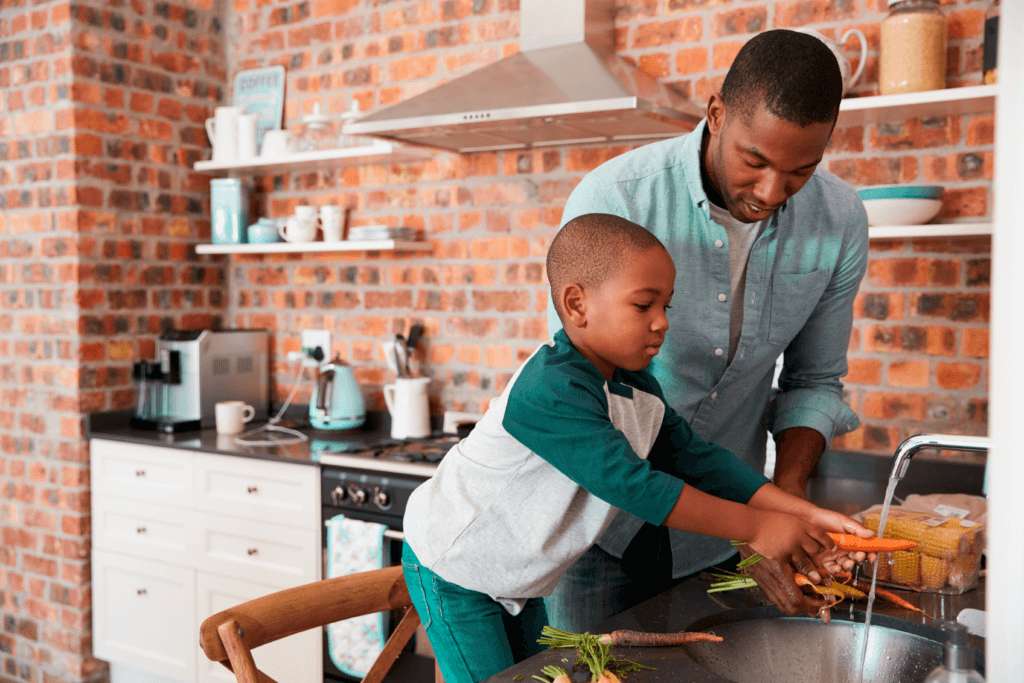 Um pai em pé ao lado de seu filho em cima de uma cadeira. Ambos estão apoiados em uma pia lavando os alimentos