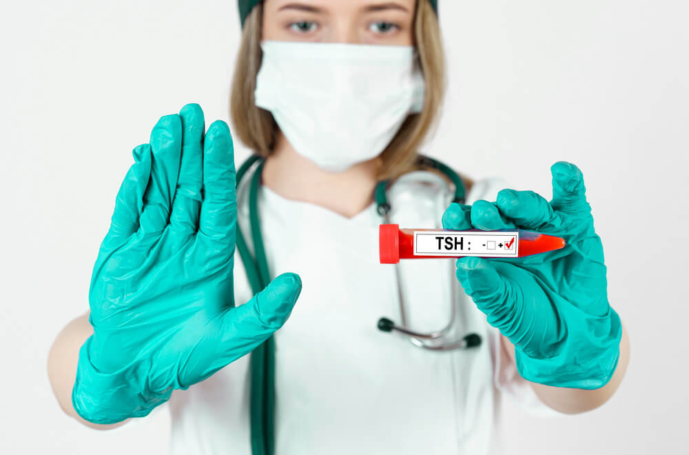 Enfermeira usando máscara, luva e estetoscópio mostrando uma ampola de sangue escrito TSH. Essa foto serve para ilustrar o post sobre o que é exame de TSH.