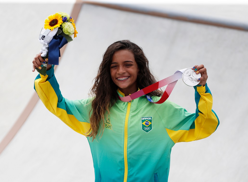 A imagem mostra : Rayssa Leal, a “Fadinha” do skate, segurando uma medalha de prata em uma mão e um buquê de girassóis na outra.
