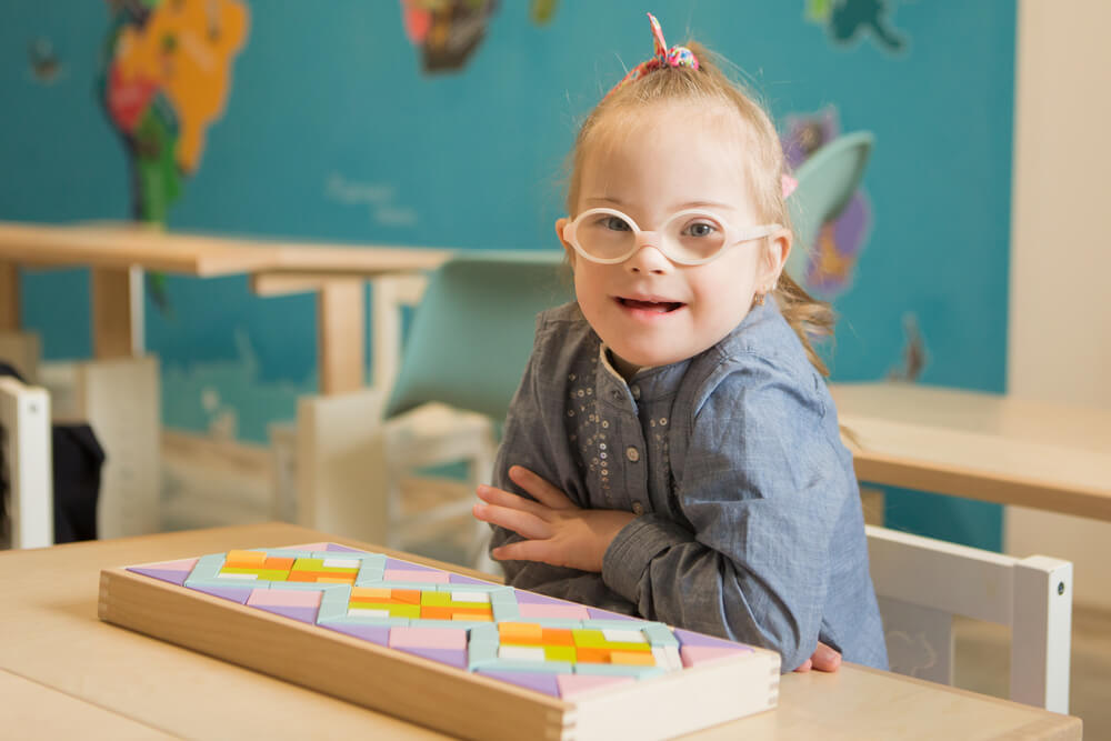 Menina com Síndrome de Down sentada em uma cadeira. Na sua frente um brinquedo em forma de mosaico. Ela está de braços cruzados e sorrindo olhando para a câmera