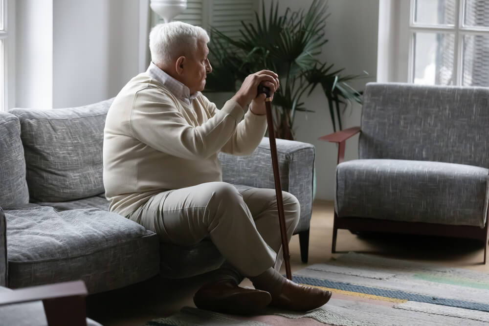 Idoso que sofre com a ataxia sentado no sofá segurando um bastão que o ajuda a andar