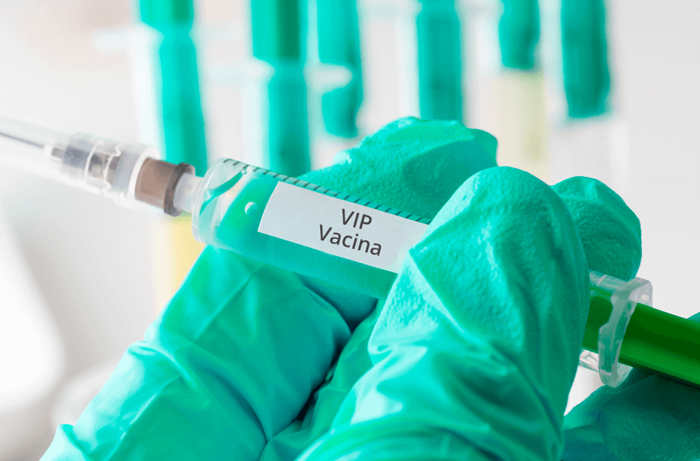 uma mão com luva verde segurando uma seringa com a descrição de vacina vip