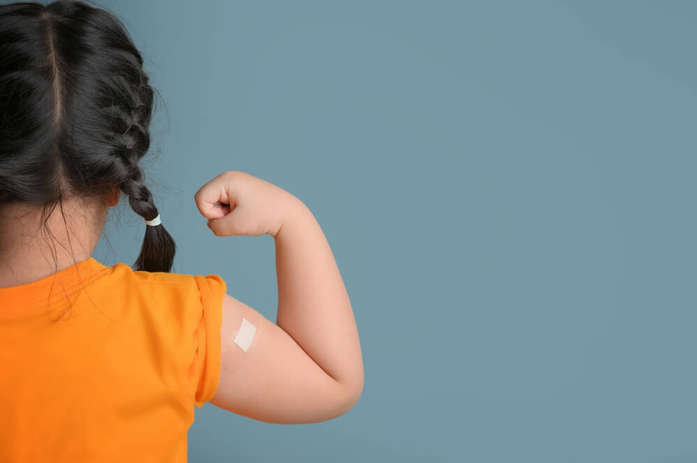 menina virada de costas e com trança no cabelo levanta o braço com um curativo após tomar a vacina meningocócica B