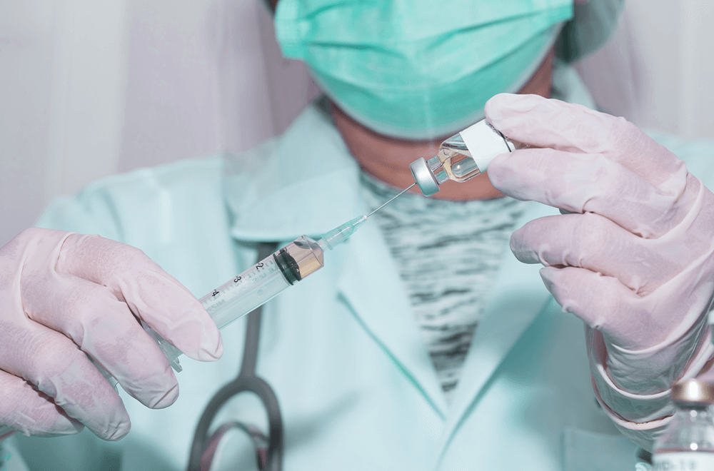 Técnica de enfermagem usando máscara facial segura uma ampola de vacina pneumocócica 13 e passa o líquido para a injeção.