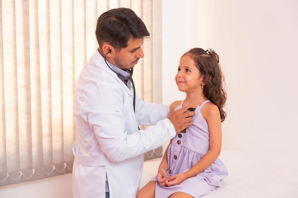 Menina sentada em cima da maca durante uma consulta com pediatra. A menina olha sorrindo para o médico enquanto ele faz uma ausculta pulmonar (com o estetoscópio)